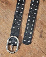 Cinturón de cuero negro con textura de reptil, decorado con tachas metálicas y ojales plateados. Cuenta con una hebilla ovalada grande de metal plateado.