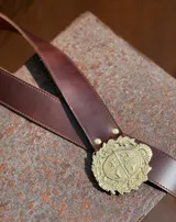 Cinturón de cuero grueso color marrón chocolate con hebilla metálica dorada con diseño de escudo rodeado de laureles.