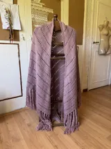 Ruana tejida a mano color malva o lila suave, con diseño de punto calado en zigzag y flecos largos en los bordes.
