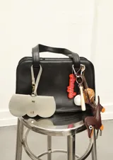 Funda para lentes de cuero sintético color gris claro con detalle de borde festoneado y cierre con broche a presión. Incluye tira para colgar de un bolso.