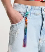 Short de jeans celeste con trabillas en la cintura, cinta de colores en las trabillas, cierre con botón y cremallera, bolsillos, desgastes y bajo deshilachado.