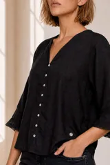 Camisa blanca de lino, con escote en V, mangas tres cuartos con detalles de encaje en los puños, cierre frontal de botones y diseño más largo en la parte trasera.