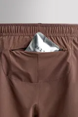 Short deportivo de entrenamiento en color marrón, confeccionado en tejido técnico ligero y elástico. Cuenta con cintura elástica ajustable con cordón, bolsillos frontales, bolsillo trasero con cierre y aberturas laterales en el bajo. Incluye un pequeño detalle de logo estampado en la pierna.