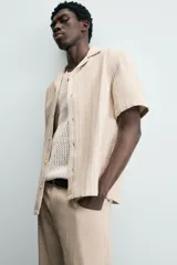 Camisa de manga corta y corte relajado, confeccionada en tejido de algodón con estructura de rayas verticales. Presenta cuello bowling, cierre frontal con botones y aberturas laterales en el bajo.