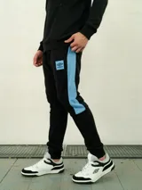 Pantalón de felpa negro con puños elásticos en los tobillos, y una franja lateral azul celeste. Incluye el logo de Umbro en la parte superior del muslo.