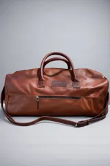Bolso de viaje tipo duffel bag de cuero marrón, con asas de mano y correa ajustable para hombro. Presenta un bolsillo exterior con cierre en la parte frontal.