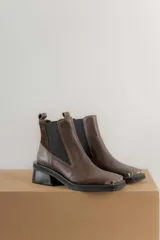 Botas tipo chelsea de cuero color marrón oscuro con acabado brillante. Presentan punta cuadrada con detalle de puntera metálica, elásticos laterales y taco cuadrado bajo.
