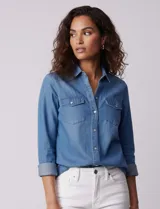 Camisa de manga larga en tela denim de color azul medio, con cuello clásico, botones frontales y dos bolsillos de parche con solapa en el pecho.