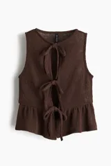 Top corto sin mangas de punto marrón oscuro, con escote redondo y abertura frontal que se cierra con tres lazos anudados. Presenta un ruedo con volado (peplum).
