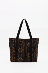 Bolso tote de algodón negro con estampado en tonos marrones y ocres, con costuras visibles en color claro. Presenta tiras de hombro negras y un adorno colgante con borlas negras y detalles dorados.