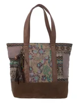 Cartera tipo tote con diseño patchwork que combina paneles de tela estampada con motivos geométricos y florales, y detalles en cuero sintético marrón. Incluye asas largas, base reforzada y un colgante decorativo de borlas y cadenas metálicas en el lateral.