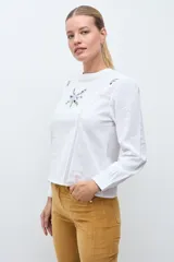 Blusa blanca de manga larga con cuello redondo, confeccionada en tela liviana con detalles de encaje vertical y bordado floral en el pecho.