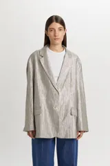 Blazer de corte oversize y estilo masculino, confeccionado en tejido con brillo metalizado y sutil patrón de rayas. Presenta solapas de muesca, cierre frontal de dos botones y bolsillos con solapa en los laterales.