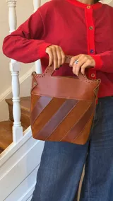 Bolso tipo matera confeccionado en cuero vacuno, con diseño estructurado y manija superior integrada. Presenta un cuerpo con franjas diagonales en gamuza de colores contrastantes (amarillo, verde y azul) sobre una base de cuero marrón, con detalles de costuras artesanales en los bordes.