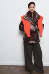Chaqueta amplia de efecto piel en color marrón oscuro, con cuello alto, manga larga y bolsillos delanteros. Presenta cintura ajustable, forro interior y cierre frontal con cremallera y botones ocultos por solapa.