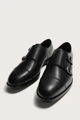 Zapato de vestir tipo monk de color negro, con detalle de doble hebilla metálica plateada en el empeine, puntera redondeada y suela con ligero tacón.
