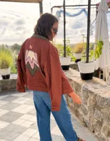 Campera bomber de jean color terracota, con corte holgado. Presenta apliques de cuero cortados y bordados a mano en la espalda, delantera y cuello, formando un diseño de montañas en tonos beige, rosa y verde militar.