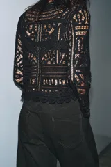 Blusa negra de manga larga confeccionada en tejido tipo crochet con intrincados patrones geométricos y florales. Presenta cuello redondo y cierre frontal con botones.