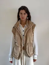 Chaleco de peluche color beige con cuello alto, cierre frontal, bolsillos laterales y detalles de etiquetas en los hombros.