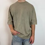 Remera de corte oversize en color verde oliva con efecto de lavado gastado. Presenta cuello redondo, mangas cortas con dobladillo ancho y costuras visibles.