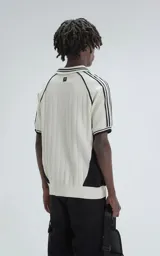 Chomba tejida de punto cerrado color off-white, con cuello polo y ribetes negros. Presenta un diseño deportivo con paneles laterales negros, número "90" y texto "Ravers" bordado en el frente.
