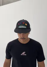 Gorro tipo gorra de béisbol color negro con visera curva. Presenta un bordado frontal de un macetero con flores de colores (rosa, azul, rojo y naranja) y el texto "OLIVER'S FLOWER GARDEN" en blanco sobre un fondo verde.