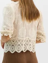 Blusa de algodón semitransparente con diseño floral bordado a tono. Presenta escote en V, cierre frontal con botones y mangas al codo ligeramente abullonadas. Incluye detalles de encaje calado estilo macramé en los puños y en el ruedo con terminaciones en ondas.