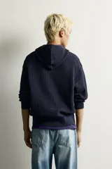 Sudadera hoodie relaxed fit color azul marino, confeccionada en tejido de algodón con estructura waffle. Cuenta con cuello con capucha, manga larga, bolsillos laterales, acabados en rib y cierre frontal con cremallera.
