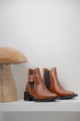 Botineta de cuero charol negro con acabado brillante, diseño de punta fina y taco cuadrado. Presenta un panel elástico lateral y una correa decorativa con hebilla metálica plateada.