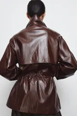 Chaqueta amplia de efecto piel en color marrón oscuro, con cuello alto, manga larga y bolsillos delanteros. Presenta cintura ajustable, forro interior y cierre frontal con cremallera y botones ocultos por solapa.