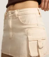 Pollera corta de sarga color marfil, de tiro medio con trabillas en la cintura y cierre frontal de botón y cremallera. Presenta bolsillos cargo laterales con solapa y hebillas decorativas, además de pespuntes en contraste.