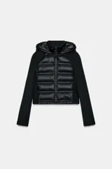 Chaqueta acolchada negra con cuello subido y capucha ajustable con cordones elásticos y stopper. Mangas largas combinadas en tejido efecto neopreno. Bolsillos laterales y cierre frontal con cremallera termosellada.