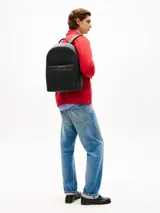 Mochila Tommy Hilfiger de color negro, confeccionada en poliuretano con paneles de texturas mixtas y diseño curvo. Cuenta con un compartimento principal con cremallera de doble cierre, funda interior para portátil, bolsillo interior con cremallera y un bolsillo exterior frontal con cierre y detalles en rojo y azul marino. Incorpora asa superior, correas ajustables y acolchadas, respaldo con panel de malla acolchado y logo metálico de la marca en la parte delantera.