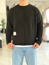 Sweater oversize de tejido de punto negro con diseño combinado, que presenta un panel frontal liso con una etiqueta decorativa blanca en el lado derecho y un panel izquierdo con textura de ochos. Tiene cuello redondo y mangas largas.