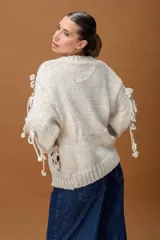 Saco de punto grueso color crudo, con diseño de calce relajado y mangas amplias. Presenta apliques tejidos con motivos florales en color terracota y detalles de tiras anudadas decorativas en toda la prenda.