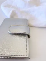 Tarjetero plateado de cuero con broche a presión. Tiene espacio para 4 tarjetas y billetes, y un bolsillo externo.