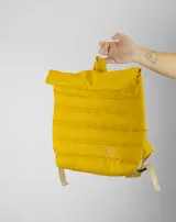 Mochila de diseño acolchado en color amarillo mostaza, con cierre superior enrollable y correas ajustables en color beige. Presenta un pequeño logo bordado en la parte frontal.