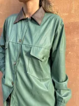 Camisa verde de cuero sintético con cuello marrón, cierre frontal con botones, dos bolsillos con solapa en el pecho y cinturón marrón.