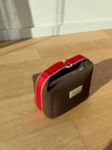 Neceser de neopreno color borgoña, con cierre superior y forro interior. Tiene un asa de mano con una franja azul y blanca.