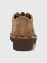 Zapato bajo de mujer color camel, estilo mocasín o derby, confeccionado en gamuza. Presenta suela gruesa de plataforma color marrón oscuro con costura visible en color claro. El sistema de ajuste es mediante cordones marrones que pasan por ojales metálicos tipo gancho.