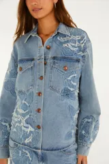 Sobrecamisa de denim azul con intrincados bordados florales en relieve, cierre frontal con botones metálicos y bolsillos delanteros con solapa.