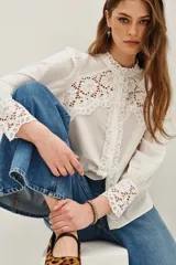Camisa blanca de algodón con cuello alto y cierre frontal de botones. Presenta detalles de encaje calado en el pecho, hombros y puños, con mangas largas abullonadas.