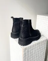 Botas cortas de cuero vacuno color negro, con diseño combinado de gamuza y cuero texturizado con efecto reptil en el talón. Presentan cierre lateral metálico, puntera reforzada y suela track dentada con costura visible.