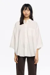 Camisa blanca de corte ancho y silueta relajada, con cuello camisero, botones frontales, manga 3/4 ancha con puño y bolsillo de plastrón en el pecho.