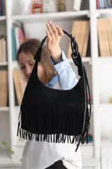 Cartera de gamuza negra con flecos largos en la parte inferior, detalle de costuras horizontales y tiras laterales ajustables.