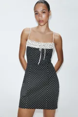 Vestido corto entallado con estampado de lunares, tirantes finos ajustables y escote redondo con detalle de frunces y lazada frontal. Presenta un diseño combinado con contraste de colores en la parte superior y cierre de cremallera oculta en la espalda.