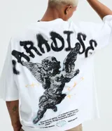 Remera oversized de algodón blanco, con cuello redondo, manga corta y gran estampa en la espalda que incluye un ángel con tatuajes, lettering "paradise" estilo graffiti y texto descriptivo en negro.