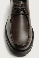 Zapato de cordones de estilo derby, color negro, con parte superior lisa, detalle de bordón en el empeine, horma redonda y suela track dentada.
