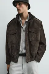 Cazadora de corte relajado confeccionada en piel con acabado de ante. Presenta cuello subido, cierre frontal de cremallera oculta por solapa con botones a presión, bolsillos de plastrón con solapa en el pecho y bolsillos de vivo en la cadera. Incluye trabillas en los hombros, mangas largas con puños abotonados y bajo con elástico.