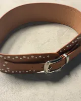 Cinturón ancho de cuero negro con doble hilera de tachas plateadas. Hebilla metálica plateada de estilo occidental y punta de cierre en forma de flecha.